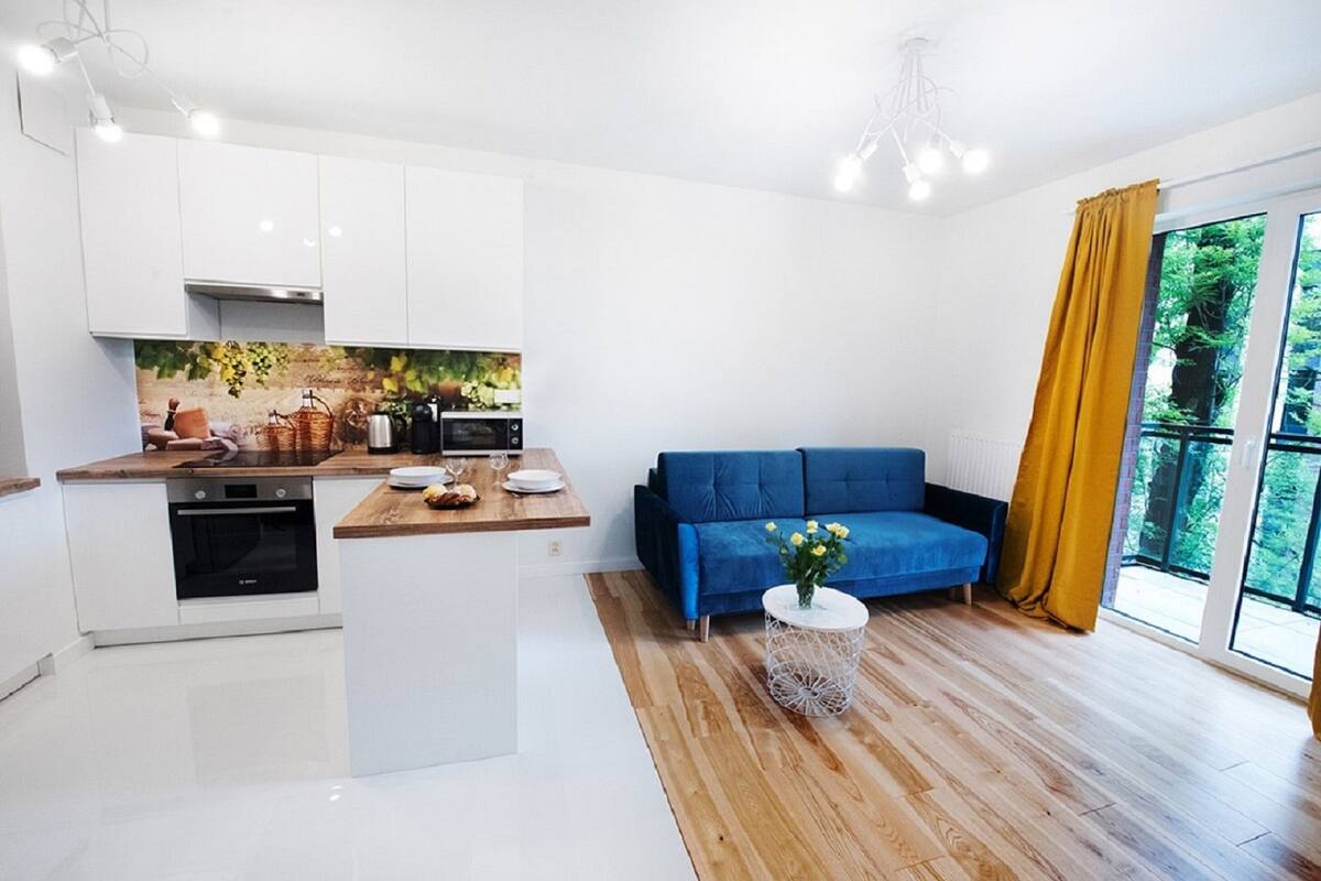 A modern kitchen with white cabinets, a wooden countertop, and a built-in oven. There is a blue sofa with a vase of flowers and a round white side table.