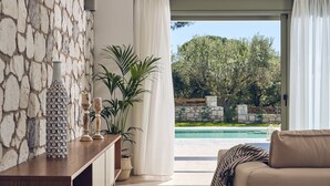 A modern living room with a stone wall, a wooden console table, a beige sofa, and a view of a swimming pool and greenery outside. - Gerakas Luxury Villas (Zakynthos)