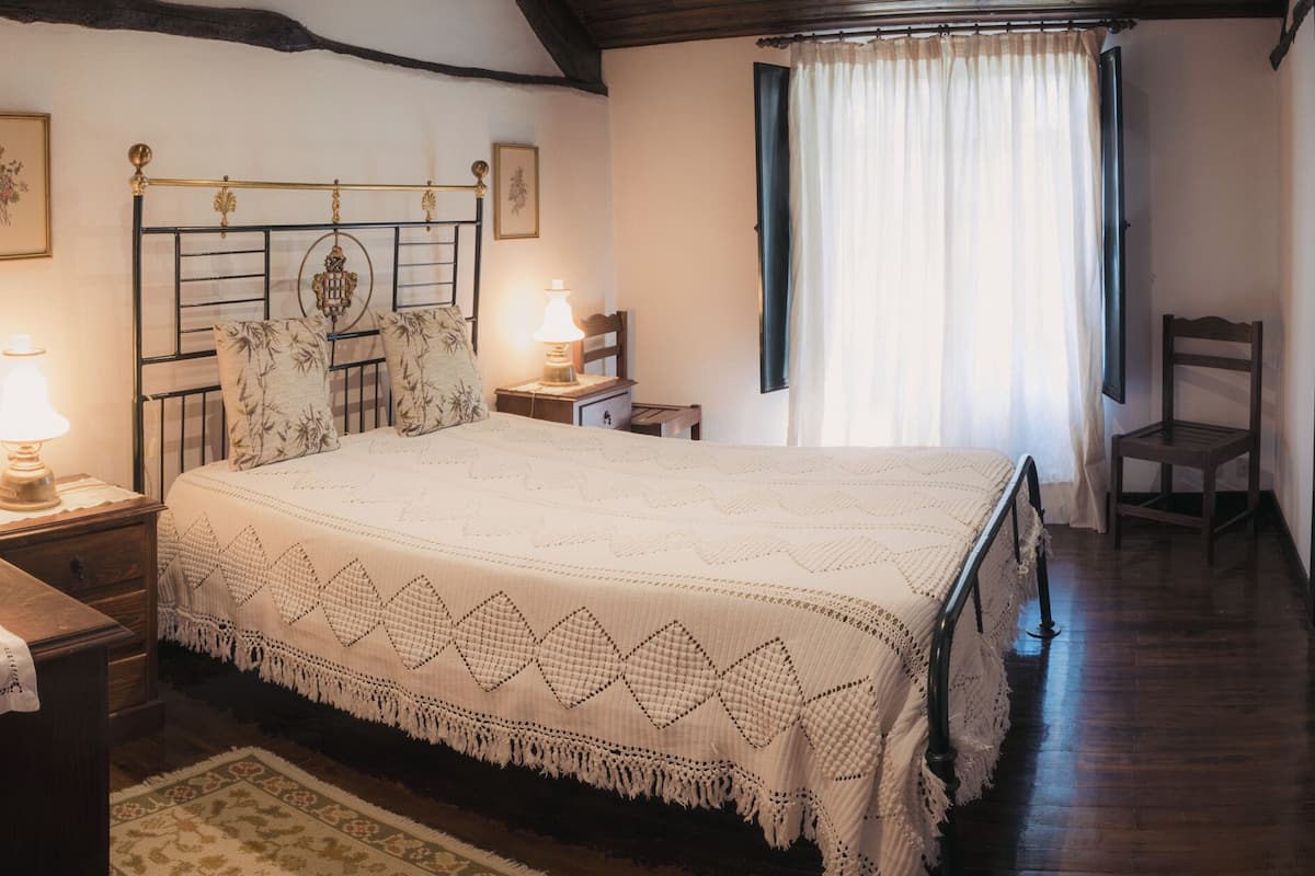 A bedroom with a bed, bedside lamps, a wooden chair, and a window with curtains.