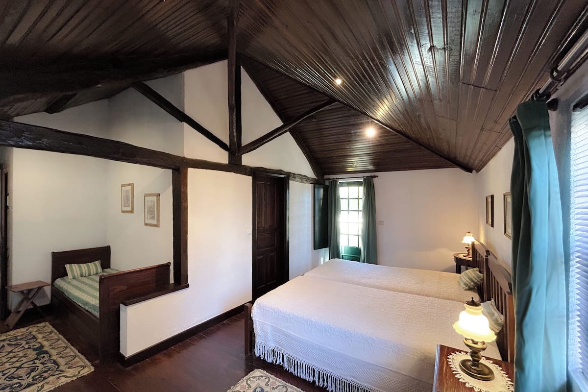 A bedroom with a large bed, a smaller bed, a window with curtains, and a wooden ceiling.