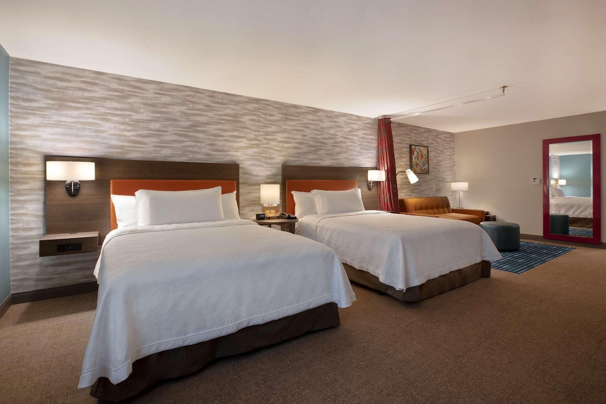 A hotel room with two beds, a nightstand with a lamp, a small table, and a mirror.