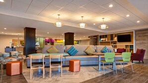 A modern hotel lobby with a sofa, chairs, a TV, and a reception desk. - Home2 Suites by Hilton Loves Park Rockford (Loves Park)