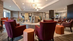 A modern hotel lobby with a seating area featuring red and orange chairs, a sofa with multiple cushions, and a dining area with tables and chairs. - Home2 Suites by Hilton Loves Park Rockford (Loves Park)