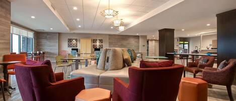 A modern hotel lobby with a seating area featuring red and orange chairs, a sofa with multiple cushions, and a dining area with tables and chairs.