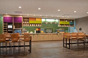 A modern cafe with wooden tables and chairs, a counter with a coffee machine, and shelves with cups and bowls. - Home2 Suites by Hilton Loves Park Rockford (Loves Park)