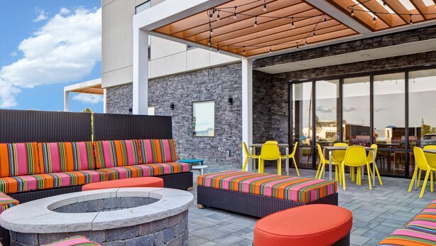 An outdoor seating area with colorful striped cushions, a stone fire pit, and yellow chairs. - Home2 Suites by Hilton Loves Park Rockford (Loves Park)