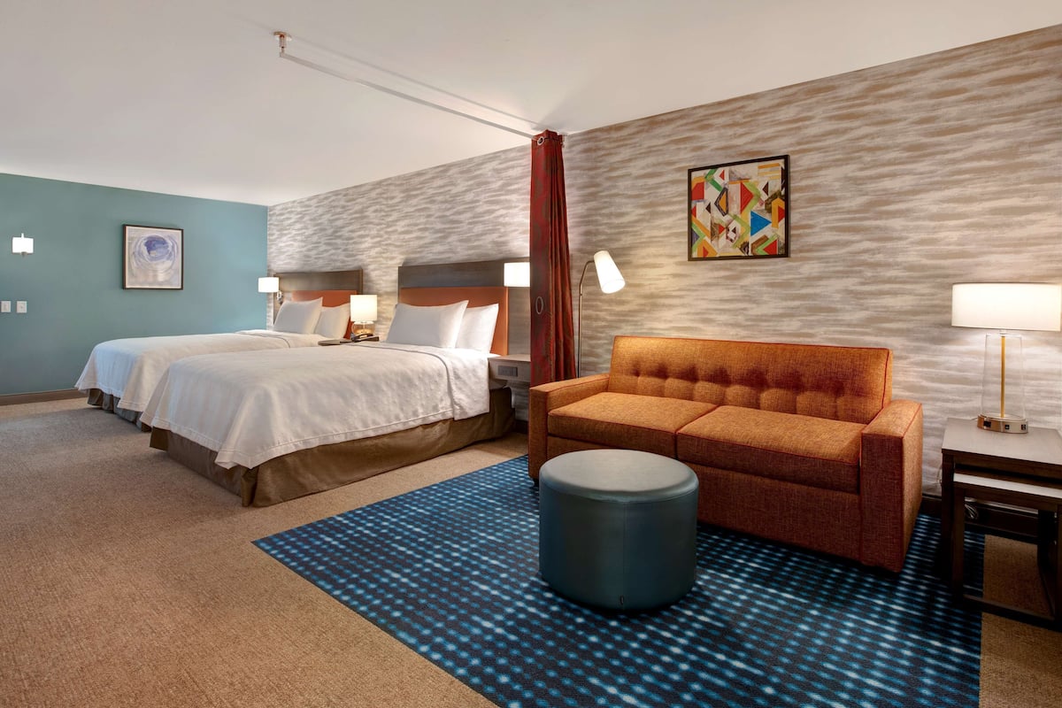 A hotel room with two beds, a sofa, a round ottoman, and a patterned rug.