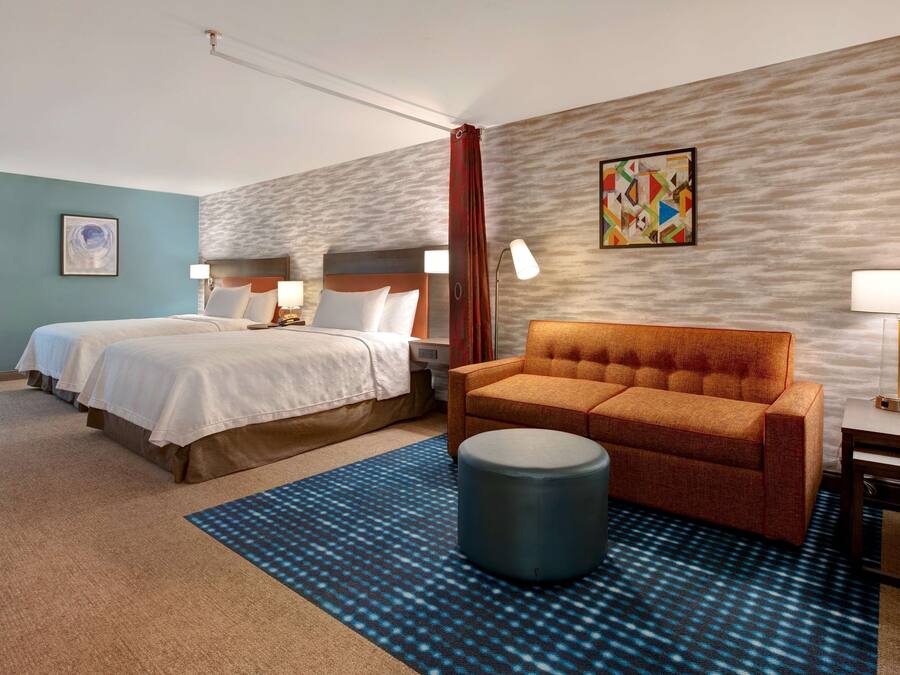 Habitación de hotel con dos camas, un sofá, un puf redondo y una alfombra estampada.