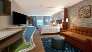 A hotel room with a desk, a bed, a sofa, a TV, and a window. - Home2 Suites by Hilton Loves Park Rockford (Loves Park)