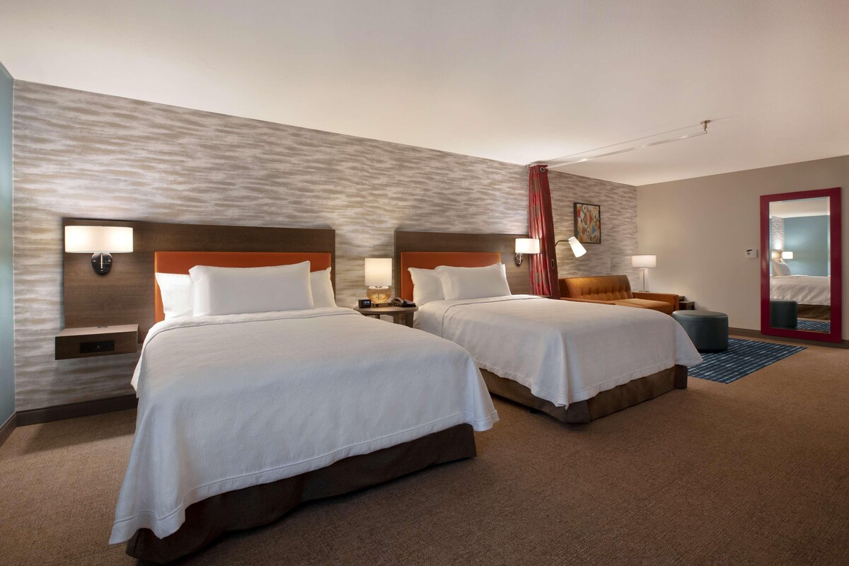 Ein Hotelzimmer mit zwei Betten, einem Nachttisch mit Lampe, einem kleinen Tisch und einem Spiegel.