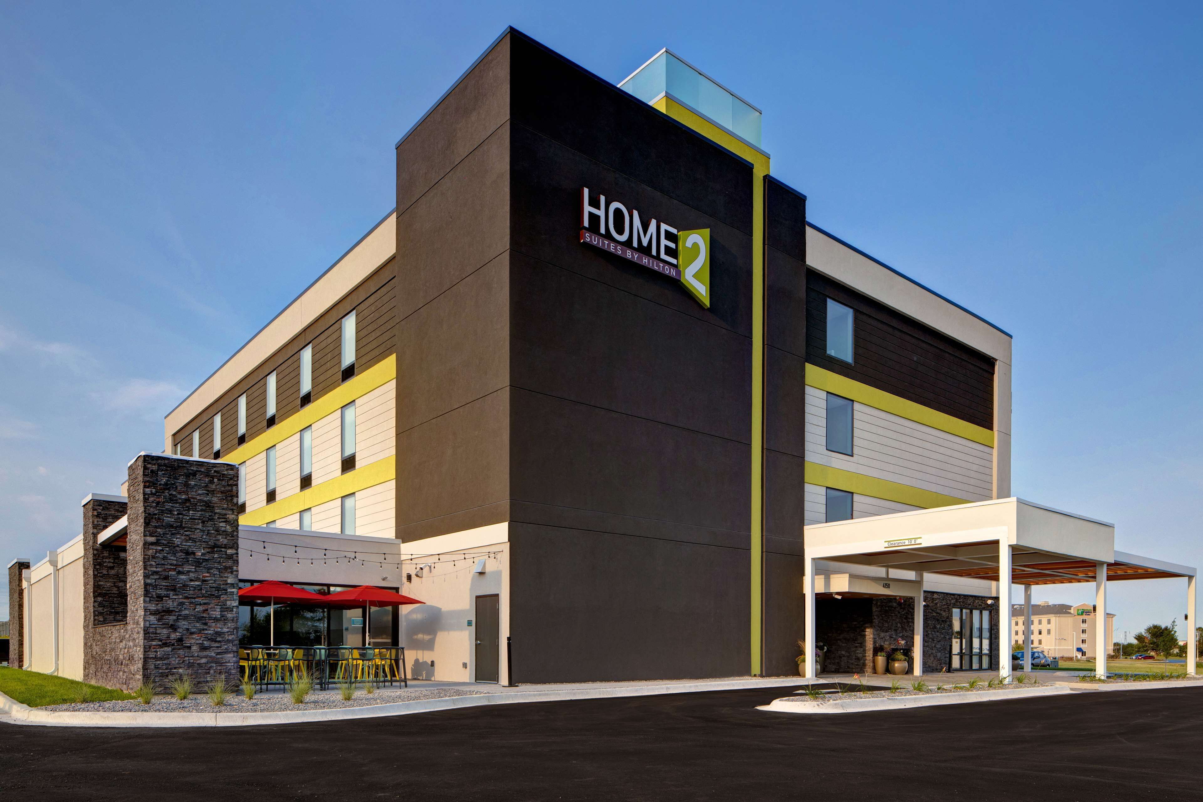 A modern hotel building with a prominent "HOME 2" sign, outdoor seating area, and a covered entrance.