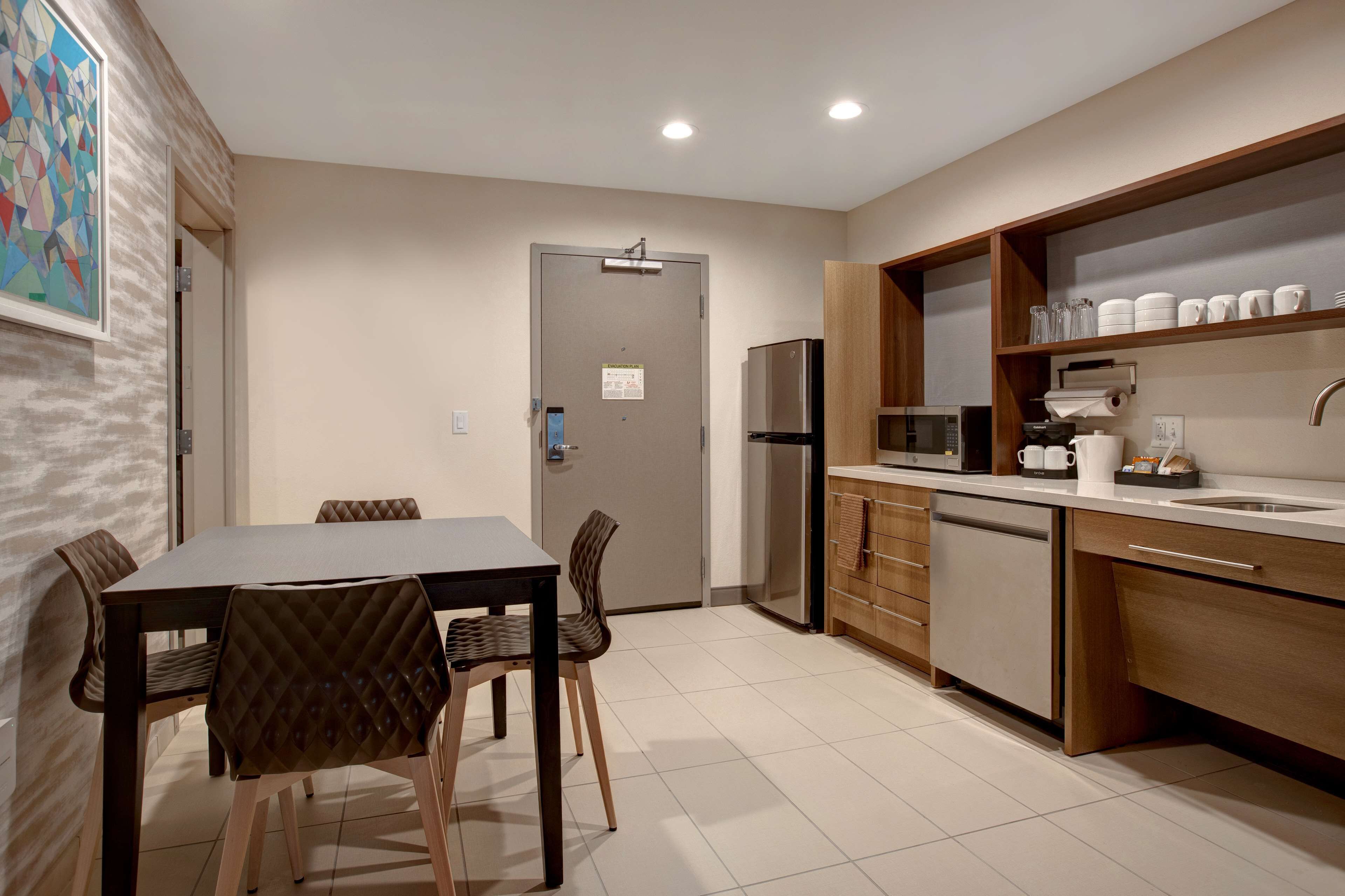 A modern kitchen with a refrigerator, microwave, and wooden cabinets. There is a dining table with chairs and a door with a sign.