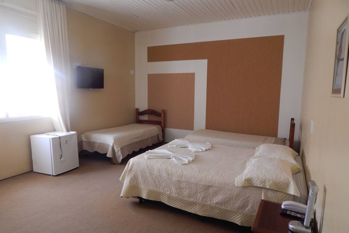 Habitación de hotel con dos camas individuales, una mesita de noche pequeña con teléfono y controles remotos, un televisor de pared y una ventana con cortinas.