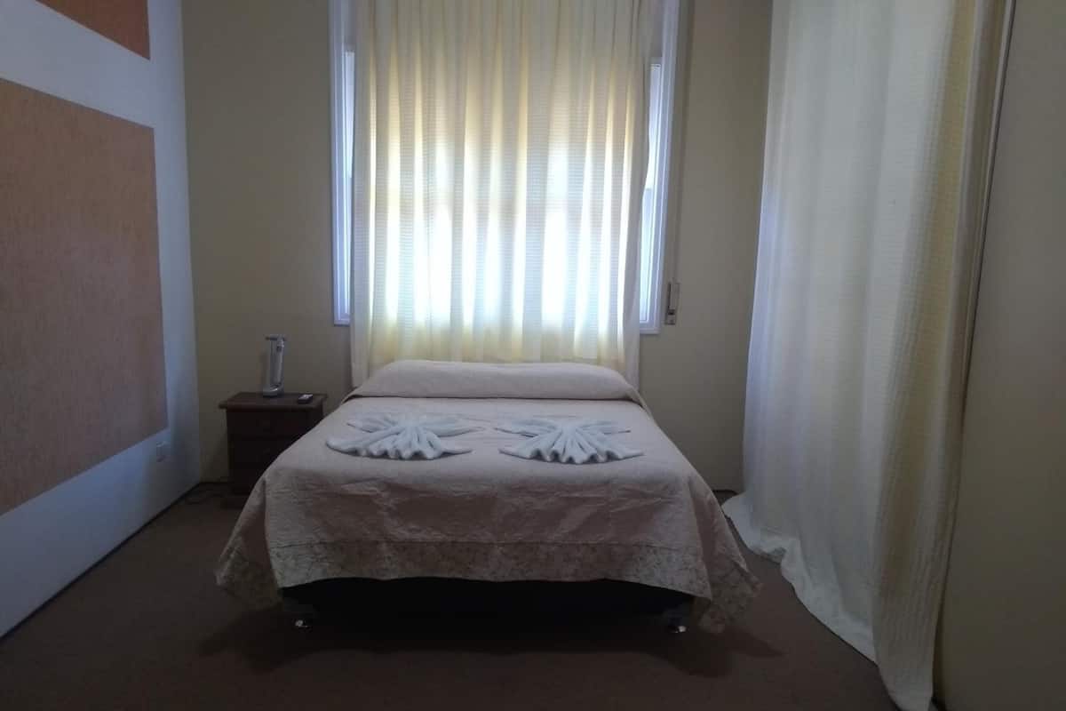 Una cama bien hecha con una colcha floral, una mesita de noche con lámpara, y una ventana con cortinas traslúcidas.