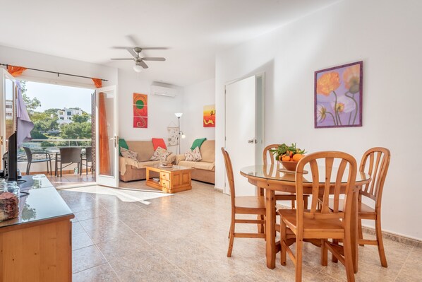 Una sala moderna con un sofá, una mesa de centro y una mesa de comedor con sillas. Cuenta con un balcón con vistas, y el ambiente se complementa con un ventilador de techo.