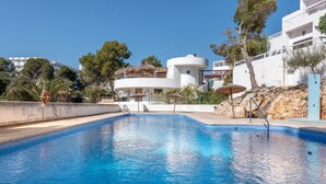 A swimming pool with a clear blue water, surrounded by a white building with a circular structure, trees, and a rocky hillside. - Air-Conditioned Holiday Apartment "Bungalow Cala Dor 27" with Sea View and Pool (Cala D'or)