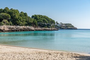 Una playa con agua turquesa cristalina, orilla de arena y un acantilado rocoso con árboles y edificios.