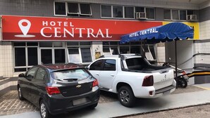 A hotel named 'Hotel Central' with a prominent red sign. There are two parked vehicles, a black car and a white SUV, in front of the hotel. - Hotel Central (Belém)