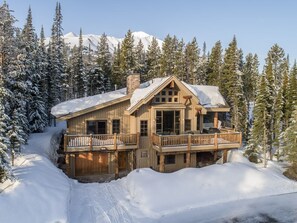 A log cabin with a balcony, surrounded by snow-covered trees and mountains in the background.
