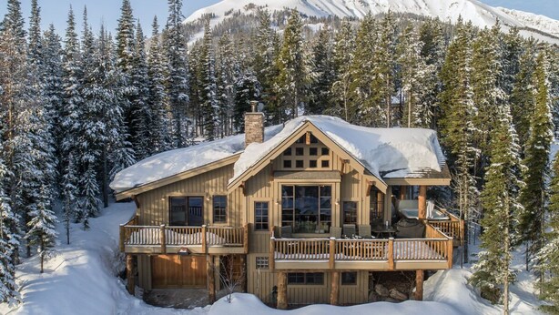 Uma cabana de tronco com varanda, cercada por árvores cobertas de neve e uma montanha ao fundo.
