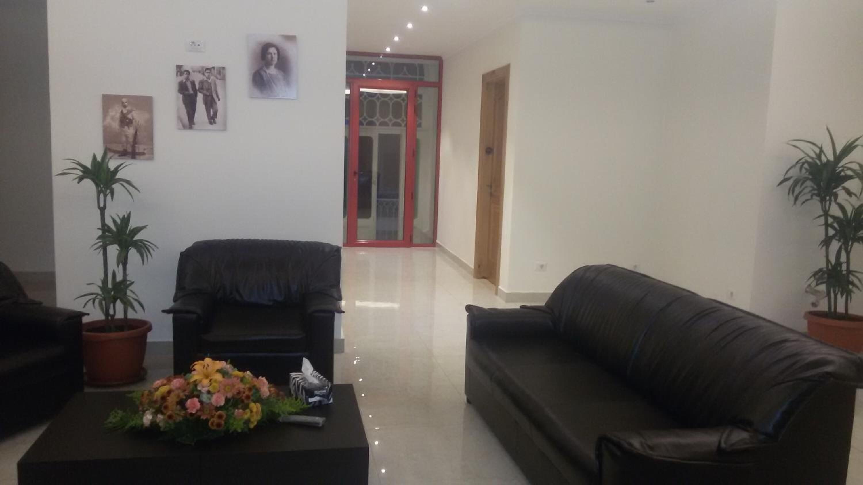 Una sala de estar con un sofá de cuero negro, un sillón negro y dos plantas en macetas. Hay un arreglo floral sobre una mesa negra.
