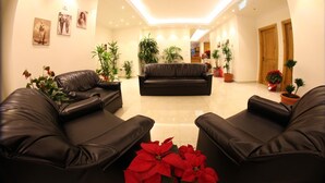 A modern living room with a black leather sofa, two armchairs, and a coffee table. There are several potted plants and a large window providing natural light. - Hôtel Saint Jean Zahle (Zahlé)