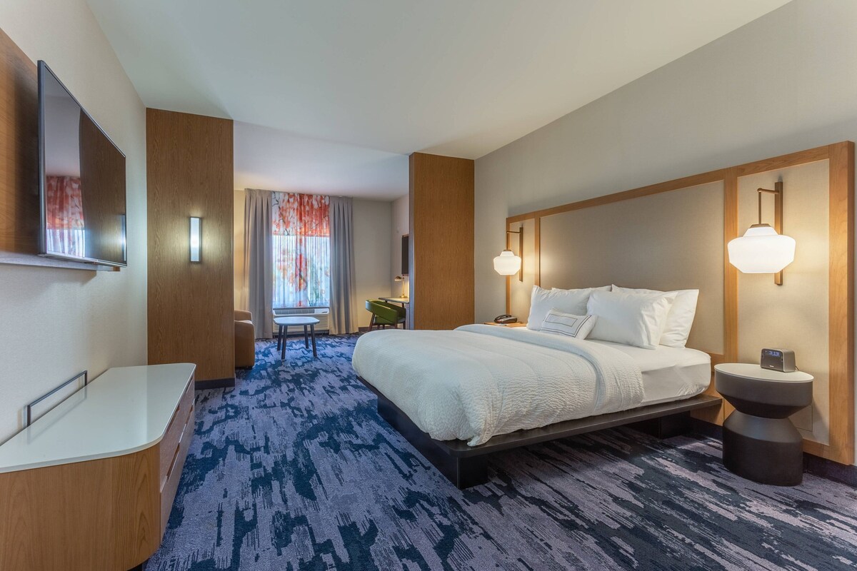 A hotel room with a large bed, a bedside table, a TV mounted on the wall, and a window with curtains.