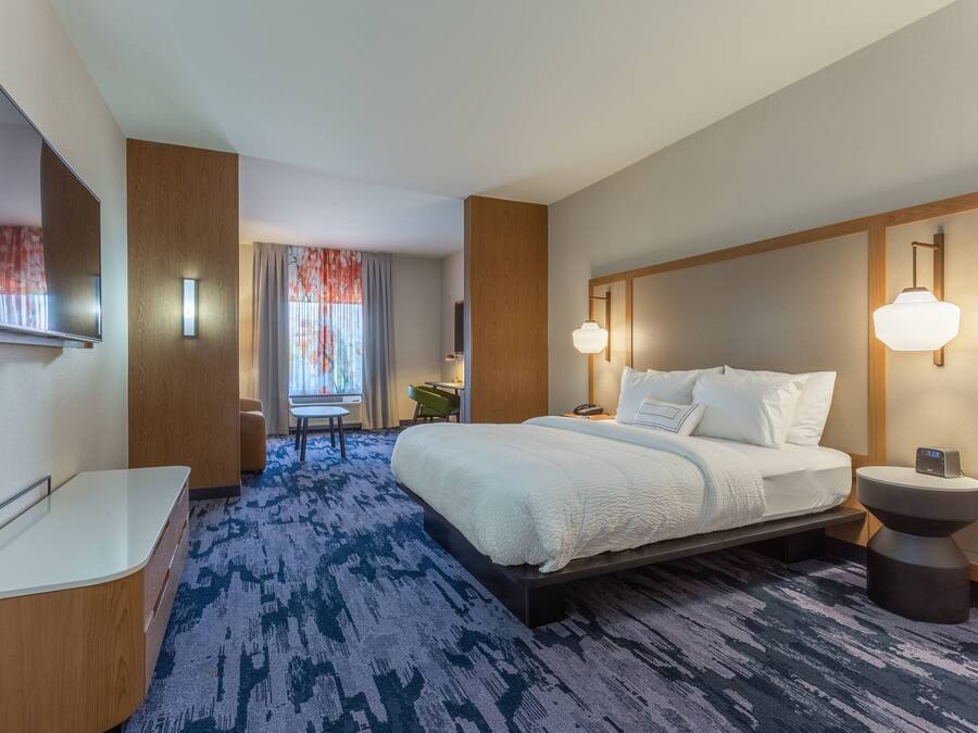 A hotel room with a large bed, a bedside table, a TV mounted on the wall, and a window with curtains.