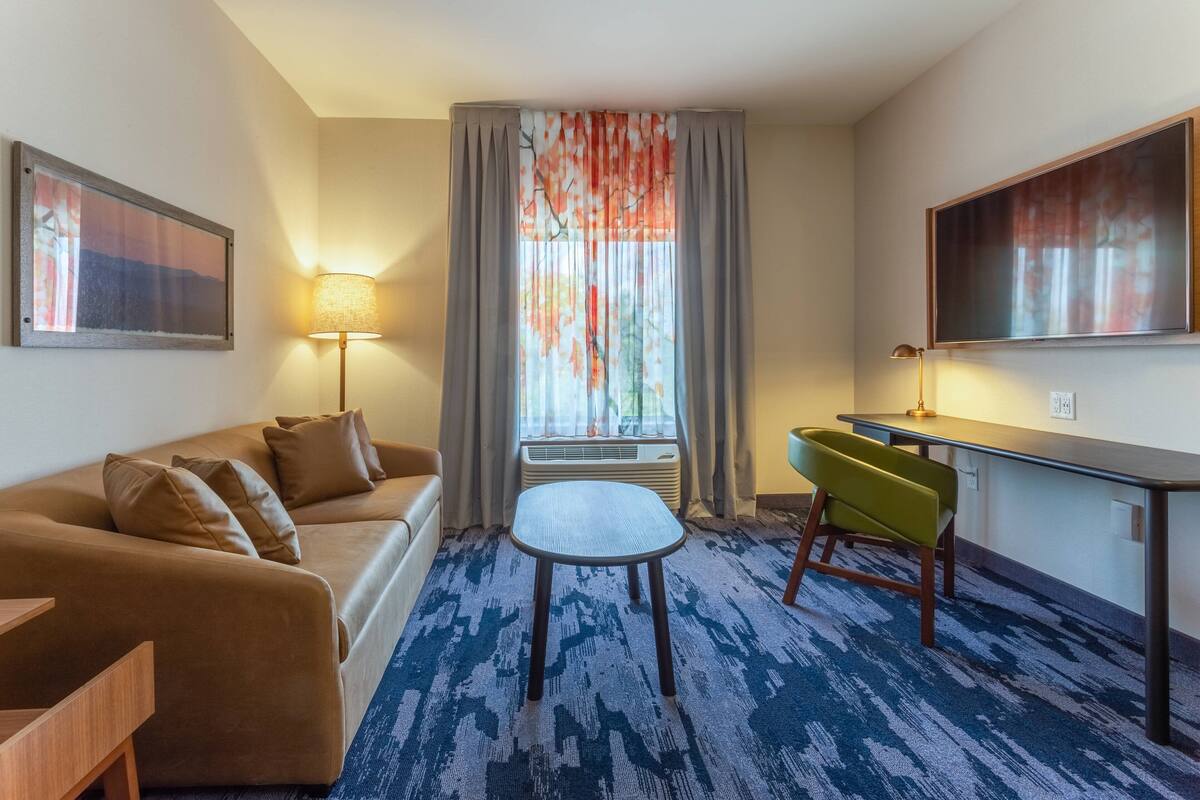 Een hotelkamer met een bank, een tafeltje, een stoel, een bureau, een lamp en een groot raam met gordijnen.