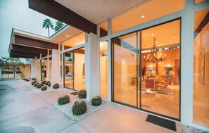 A modern house with large glass doors, a patio with cacti, and a chandelier visible inside. - Famous Pink Door | Pool | Billiard Room (Palm Springs)