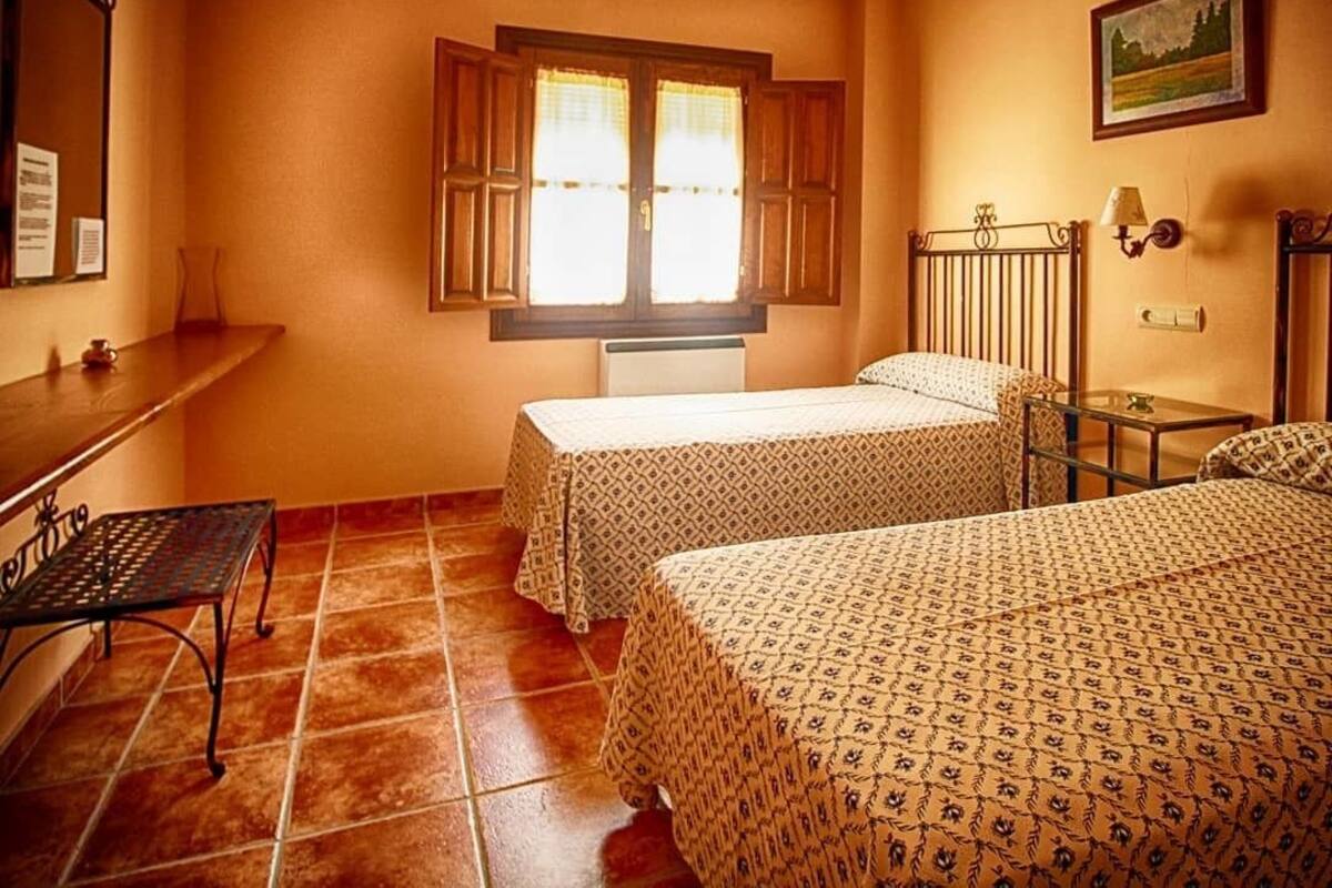 A room with two beds, a window with wooden shutters, a radiator, a small table, and a wall-mounted lamp.