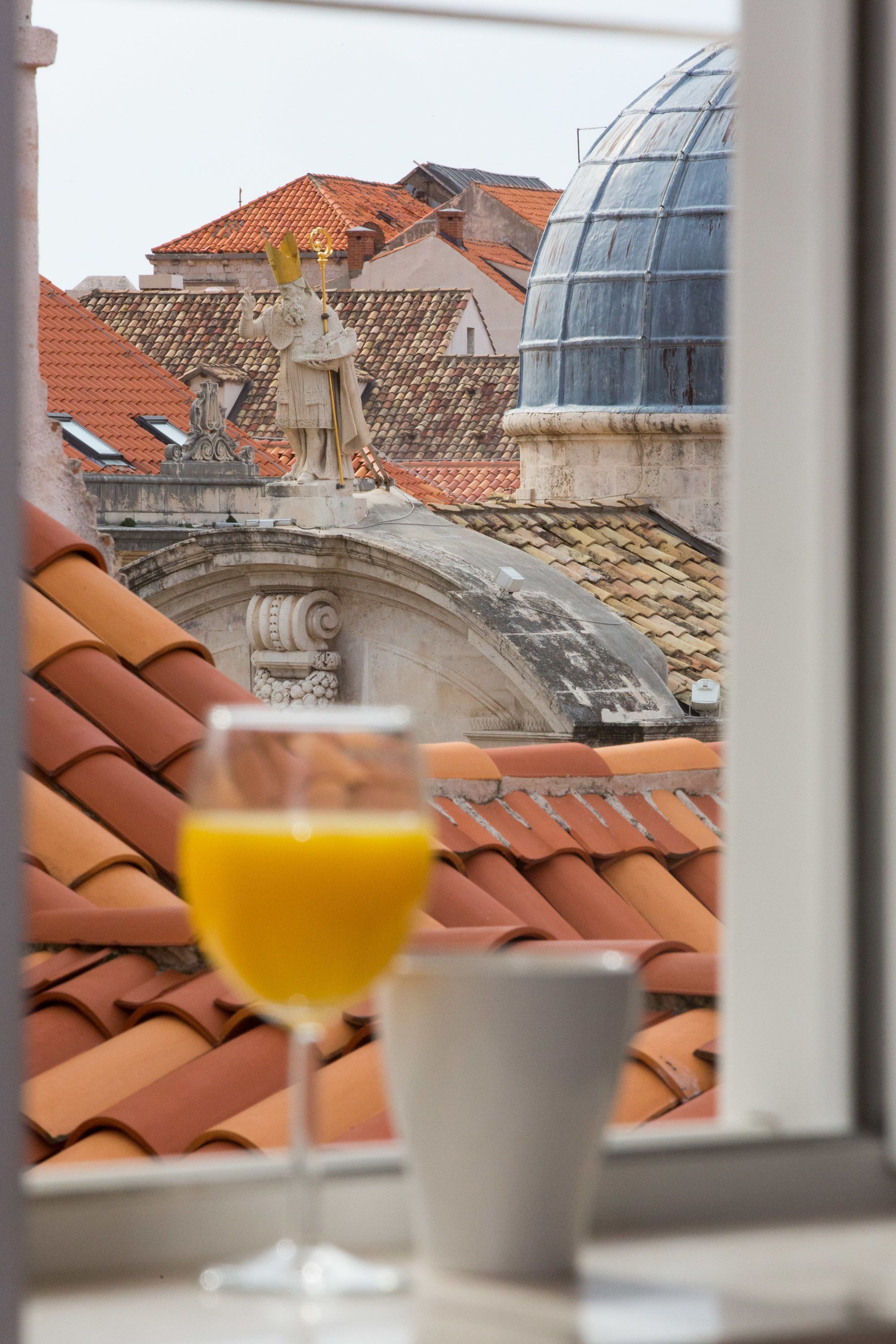 Un verre de jus d’orange sur une table, avec vue sur des toits en tuiles rouges et une statue.