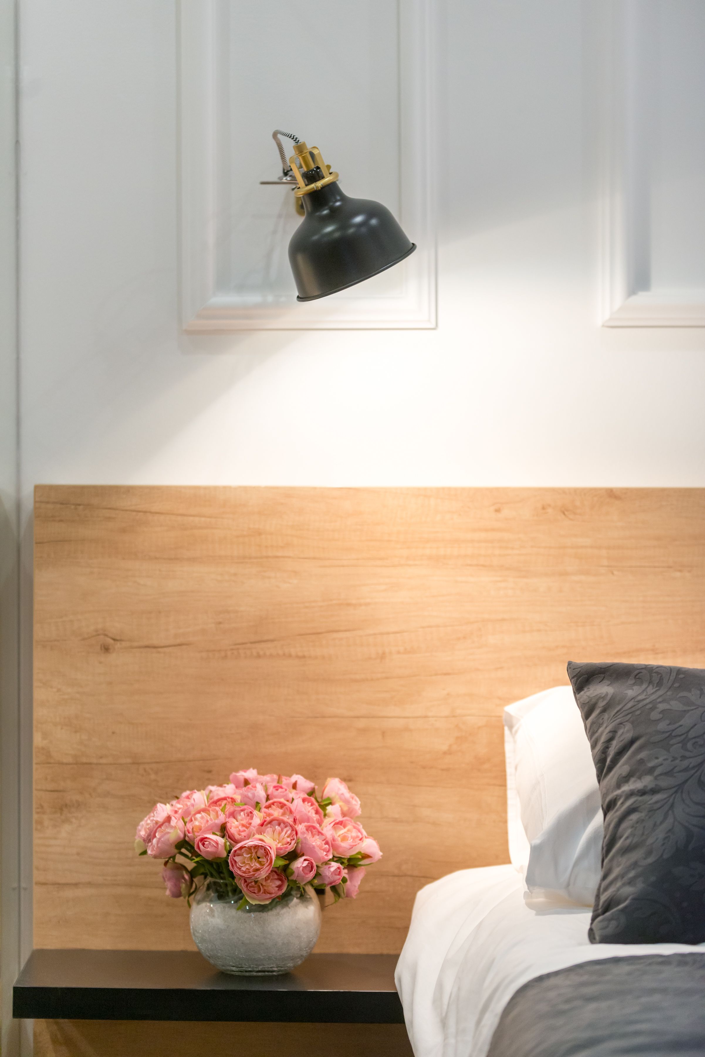 Un dormitorio ordenado con cabecera de madera, una mesita de noche con un jarrón de flores rosas y una lámpara de escritorio negra en la pared.