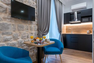 A modern kitchen with a flat-screen TV, a microwave, and a coffee maker. There is a breakfast table with fruits, a chair, and a window with curtains.