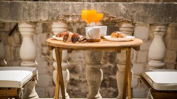 Una mesa de madera con croissants, un vaso de jugo de naranja y tazas blancas en un balcón con barandas de piedra.