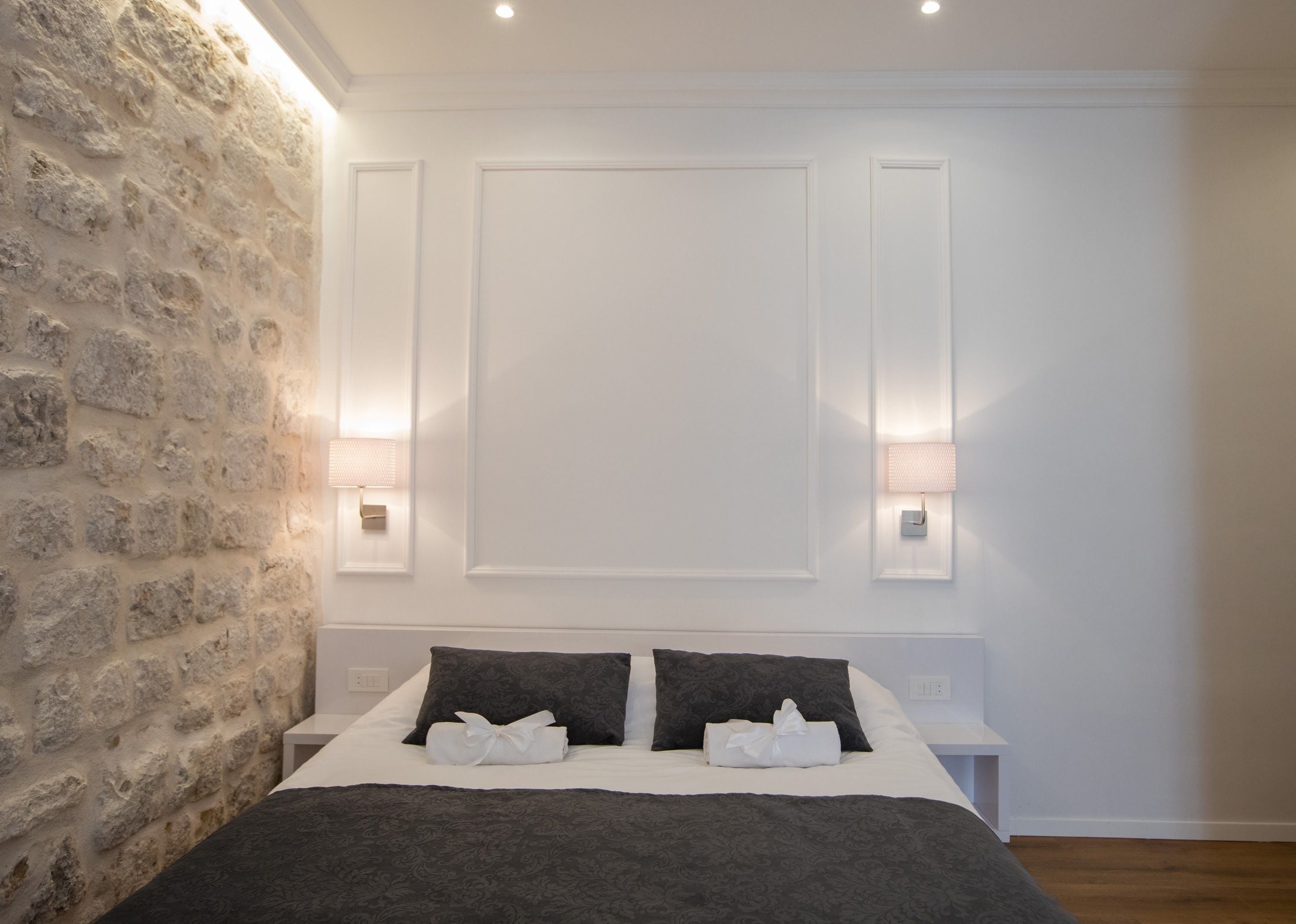 Un dormitorio con pared de piedra, cama con ropa oscura, dos lámparas de noche y un panel de techo blanco.