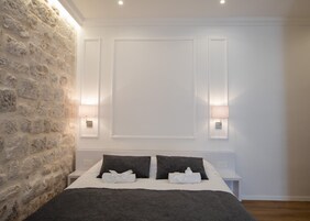 Quarto com parede de pedra, cama com roupa de cama escura, duas luminárias de cabeceira e painel de teto branco.