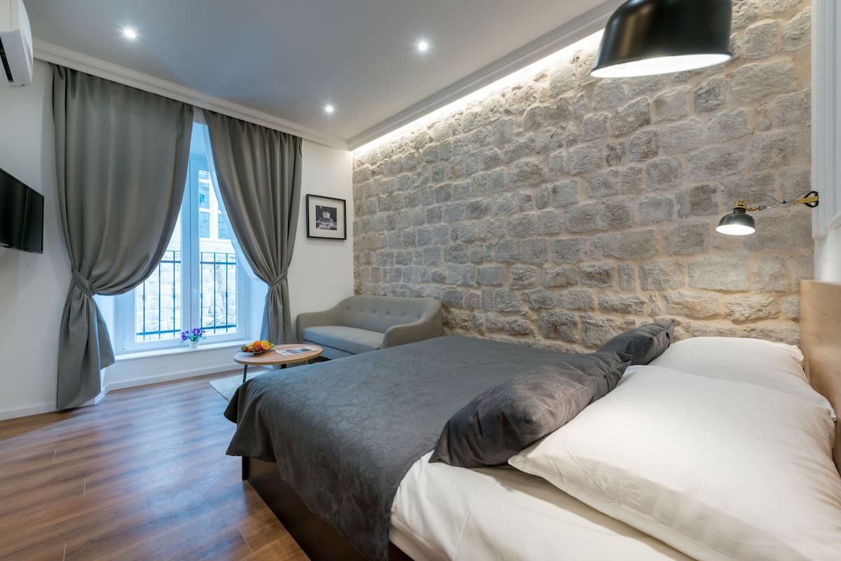 A bedroom with a stone wall, a bed with grey bedding, a grey sofa, and a small round table with a vase of flowers.