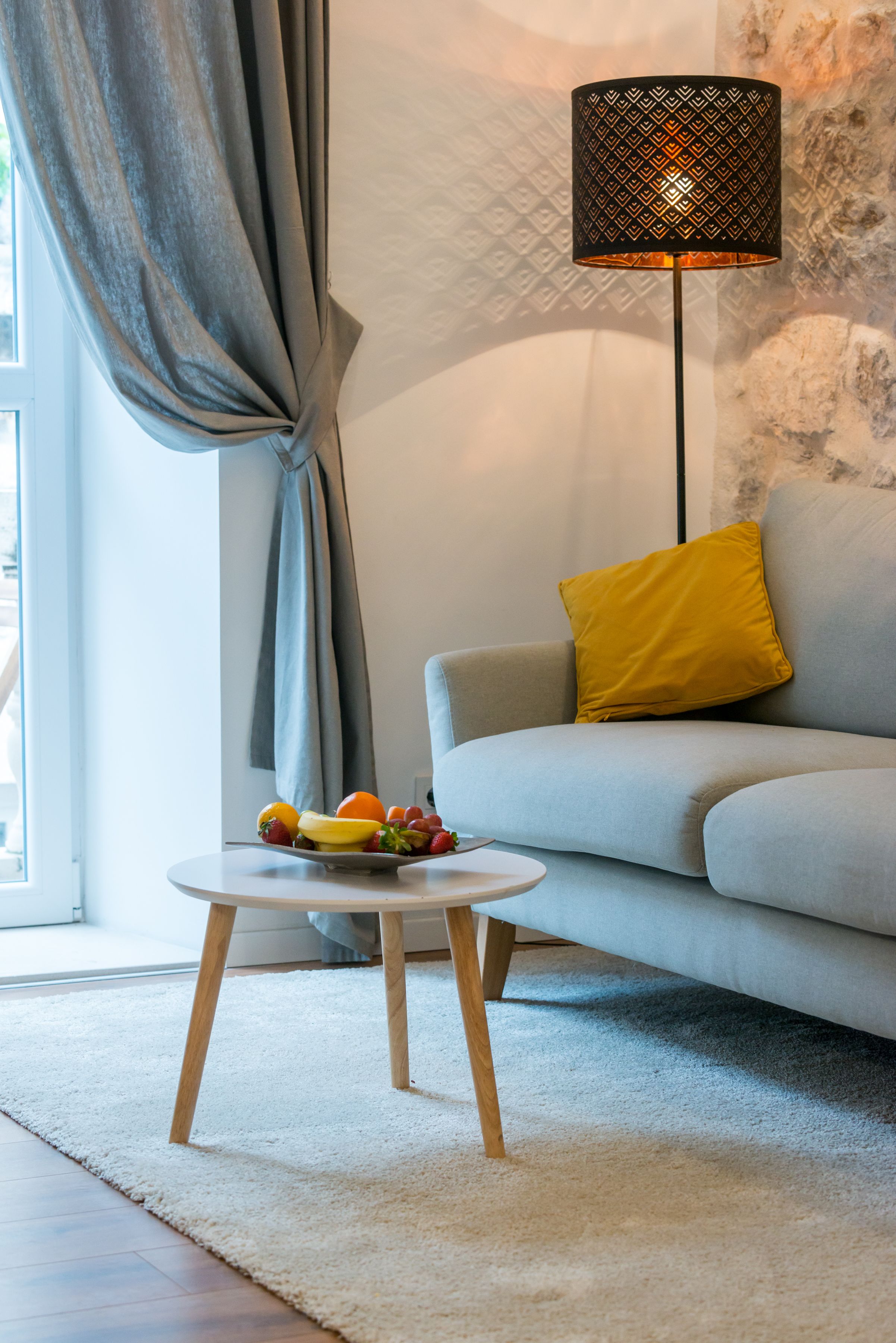 A modern living room with a grey sofa, a yellow pillow, a round white table with fruits, and a patterned floor lamp.