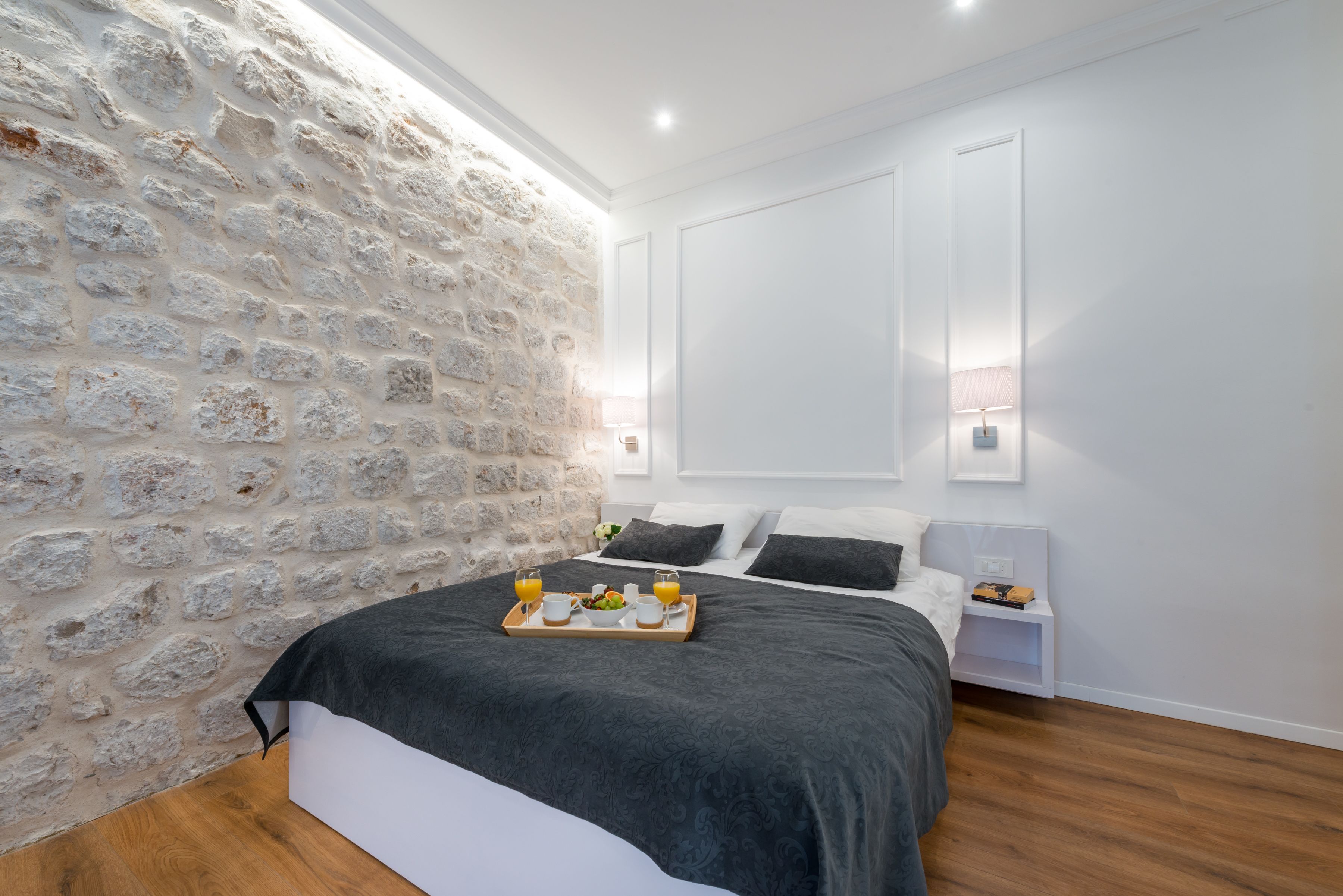 A bedroom with a stone wall, a bed with a tray of breakfast, and bedside tables with lamps.
