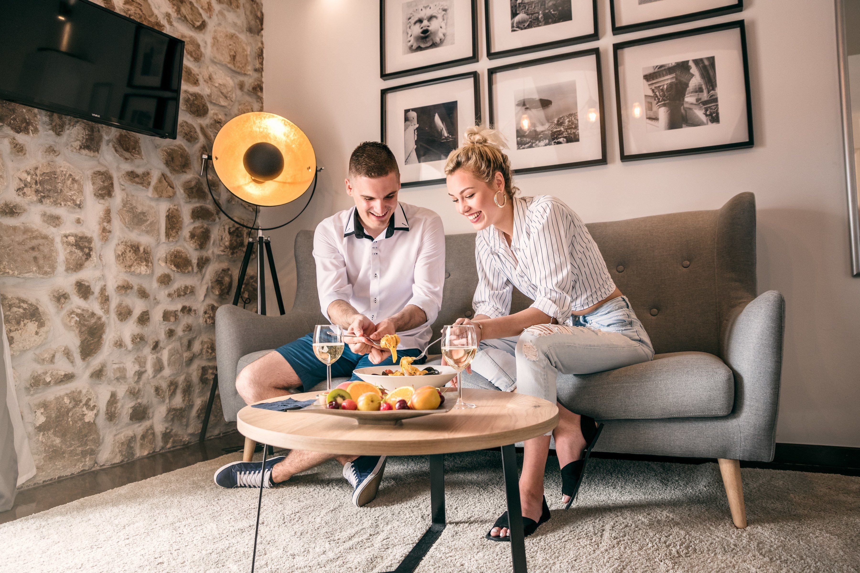 A couple dining at a round table with a fruit platter, in a room with a stone wall and framed pictures on the wall.