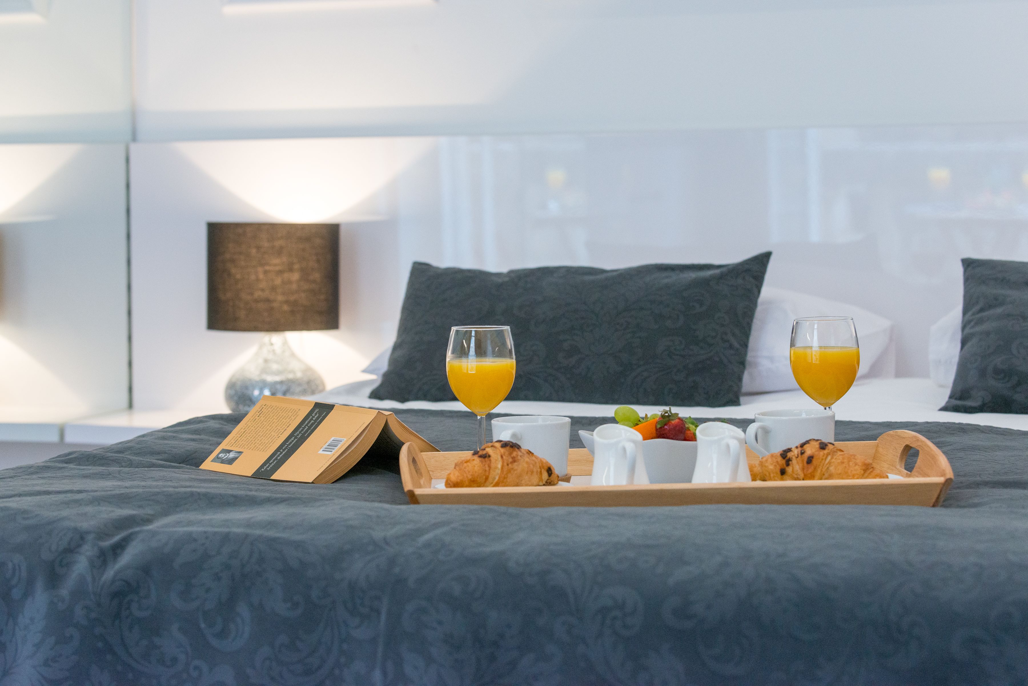 A neatly made breakfast tray on a bed with a book, two glasses of orange juice, croissants, and fresh fruit.