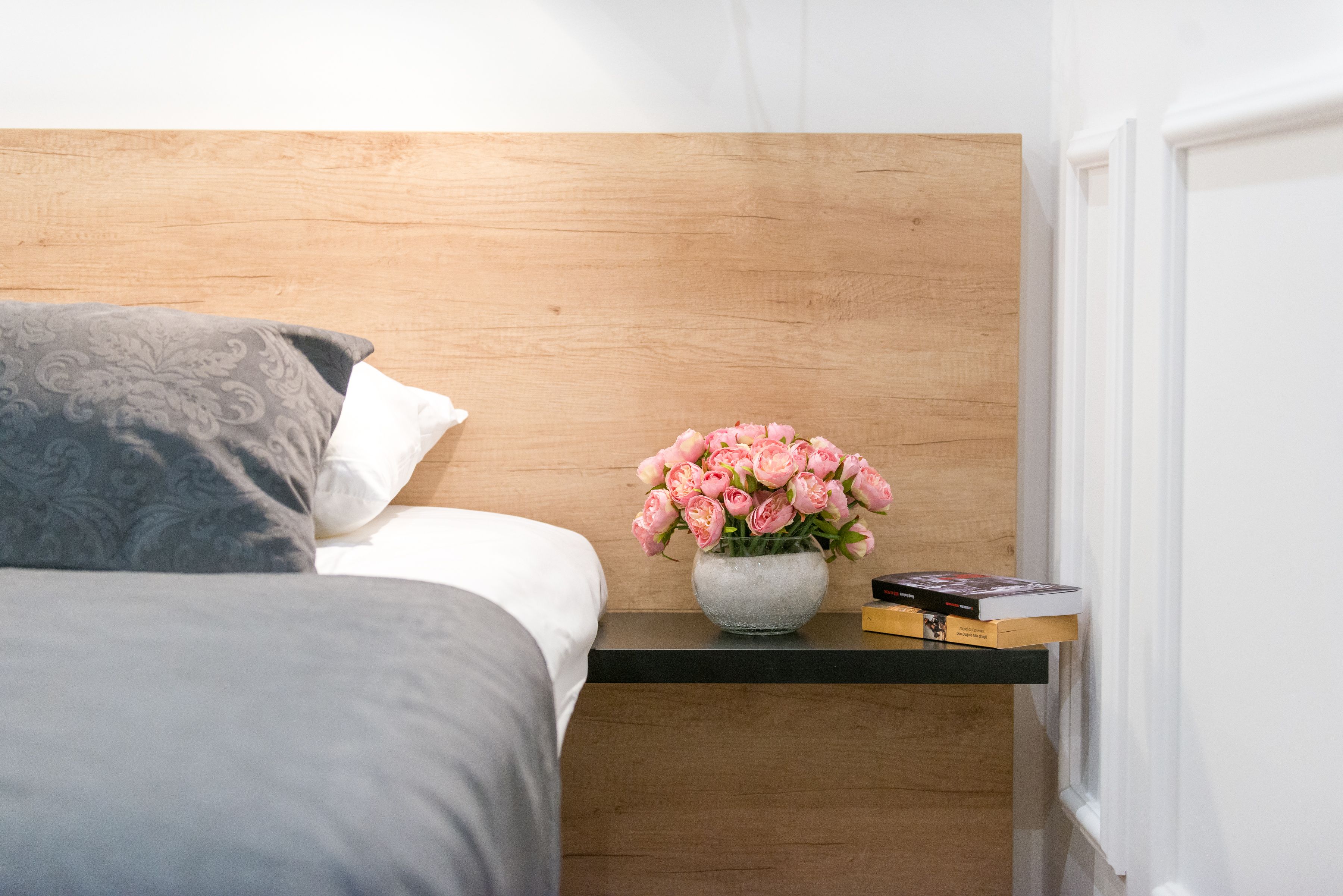 Een netjes opgemaakt bed met een grijs gestreept kussen, een klein houten nachtkastje met een vaas roze bloemen en een boek.