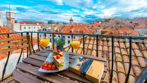 A balcony with a table set for a meal, including a sandwich, tomatoes, and a drink, overlooking a cityscape with red-tiled roofs and historic buildings. - Scalini Palace (Dubrovnik)