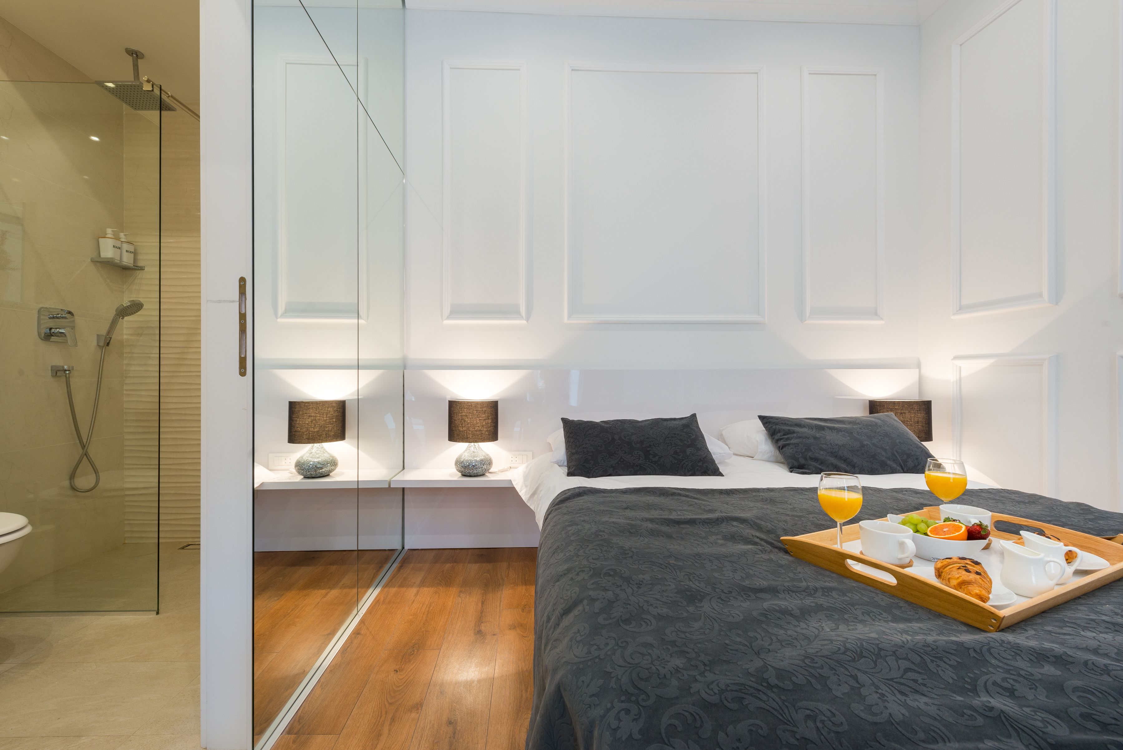A modern hotel room with a glass shower enclosure, a bed with a dark patterned bedspread, bedside lamps, and a breakfast tray with orange juice, croissants, and fruit.