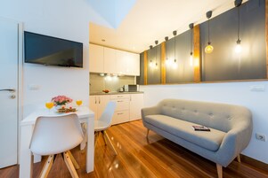 Cozinha moderna com televisão de tela plana, área de refeições com mesa e cadeiras brancas e um sofá cinzento.