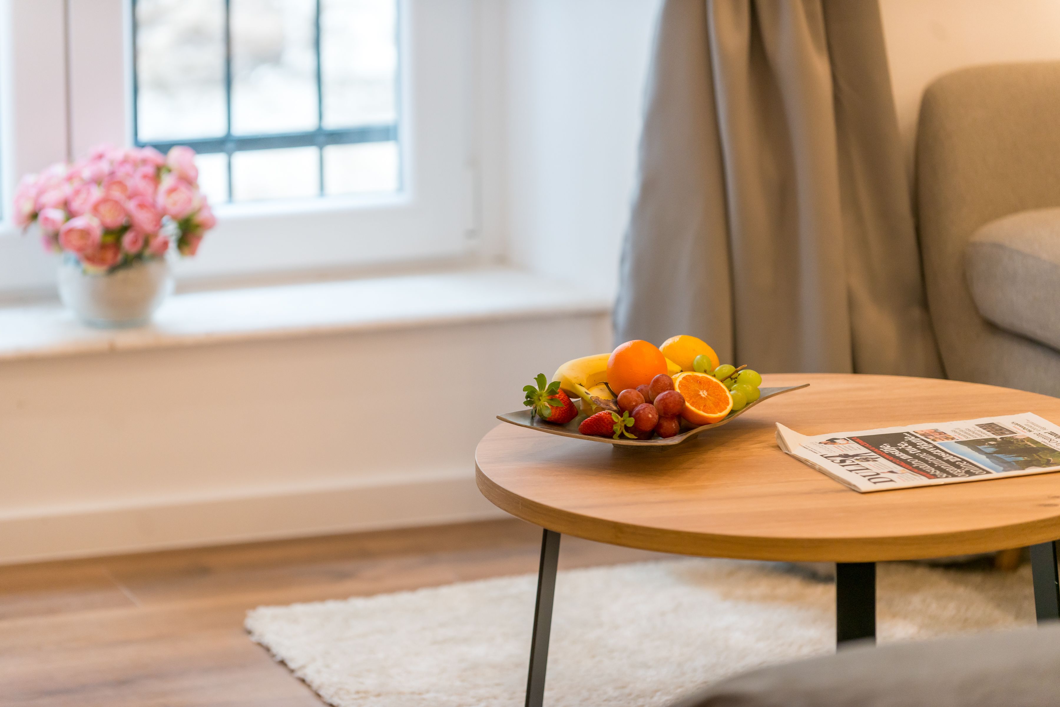 A round wooden table with a newspaper and a bowl of fruit.