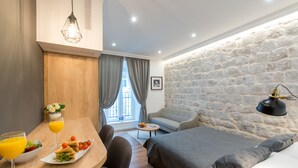 A modern hotel room with a stone wall, a bed, a sofa, a dining area with a table and chairs, and a window with curtains. - Scalini Palace (Dubrovnik)
