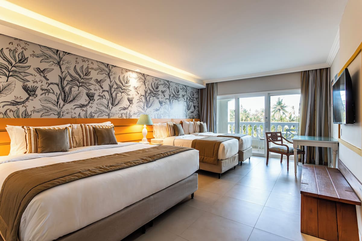 Habitación de hotel con dos camas, un escritorio, una silla y un ventanal con vistas a palmeras.
