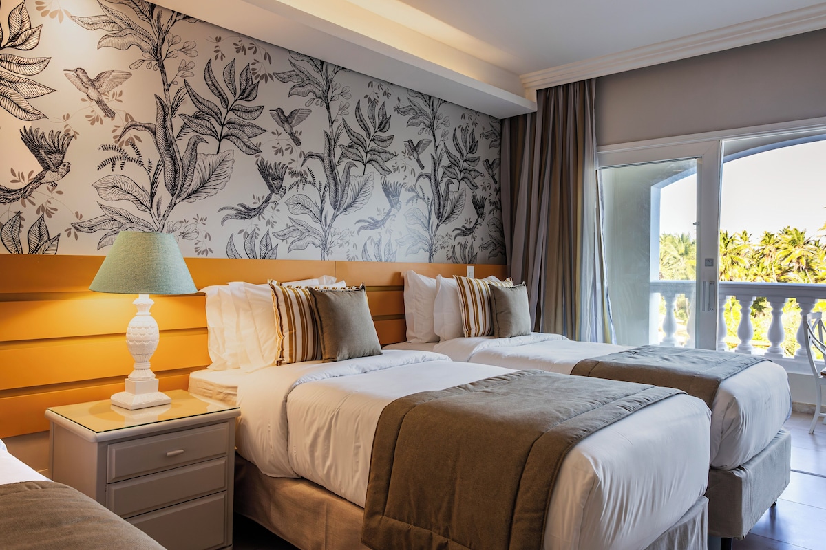 Une chambre d’hôtel avec deux lits, une table de chevet avec une lampe, un papier peint à motifs floraux et un balcon avec vue.